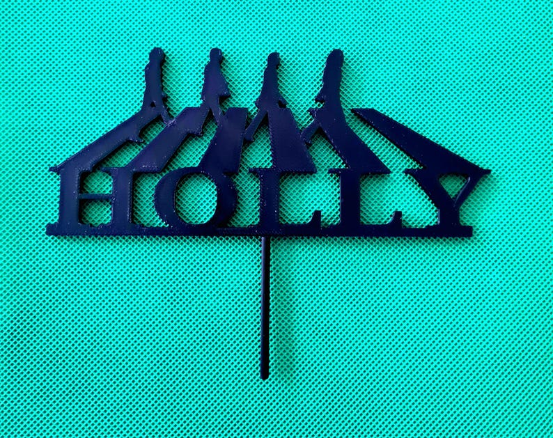 May include: A dark blue cake topper with the name "HOLLY" cut out in a stylized font. The topper features a silhouette of a circus tent with three figures on top. The topper is on a green mesh background.