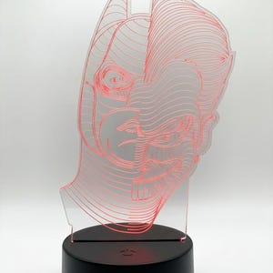 May include: A red-lined Batman head design is illuminated by a black base. The acrylic art piece features a detailed outline of the superhero's mask and cowl. The base has a power button and a small light.