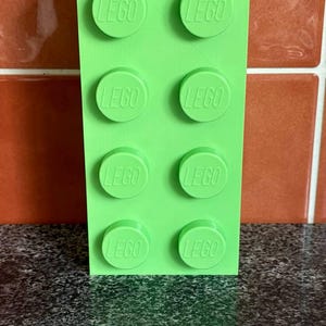 May include: A bright green Lego brick with eight circular studs, each embossed with the word "LEGO". The toy is rectangular and stands upright against a textured background.