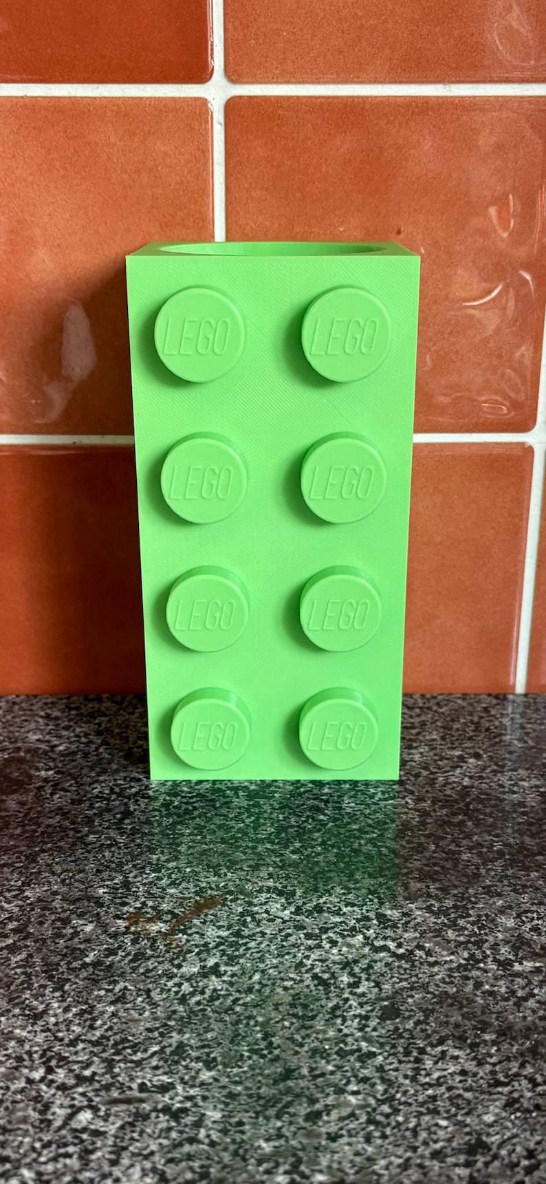 May include: A bright green Lego brick with eight circular studs, each embossed with the word "LEGO". The toy is rectangular and stands upright against a textured background.