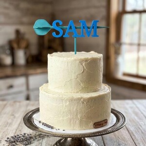 Puede incluir: Un pastel de dos niveles con glaseado blanco sobre un soporte plateado. Un adorno azul para pastel deletrea "SAM" con un dise&ntilde;o de dardo. El pastel est&aacute; decorado con glaseado texturizado.