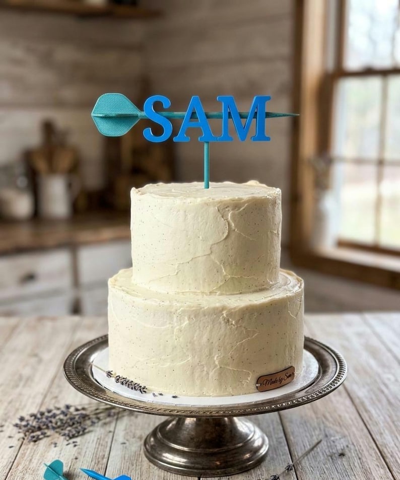 Puede incluir: Un pastel de dos niveles con glaseado blanco sobre un soporte plateado. Un adorno azul para pastel deletrea "SAM" con un dise&ntilde;o de dardo. El pastel est&aacute; decorado con glaseado texturizado.