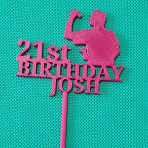 May include: A pink cake topper with the text "21st BIRTHDAY JOSH" and a silhouette of a person flexing their bicep. The topper is made of a solid material and has a long, thin stem for insertion into a cake.