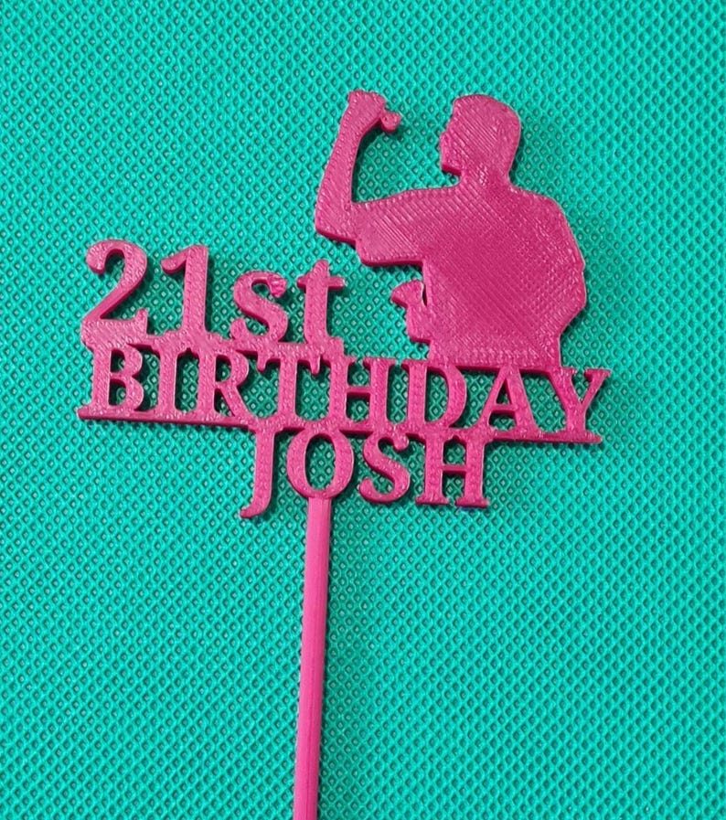 May include: A pink cake topper with the text "21st BIRTHDAY JOSH" and a silhouette of a person flexing their bicep. The topper is made of a solid material and has a long, thin stem for insertion into a cake.