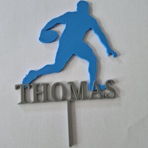 May include: A blue 3D-printed silhouette of a basketball player in motion, positioned above the name THOMAS in grey capital letters. The entire piece is attached to a single vertical grey stake, designed as a decorative cake topper.