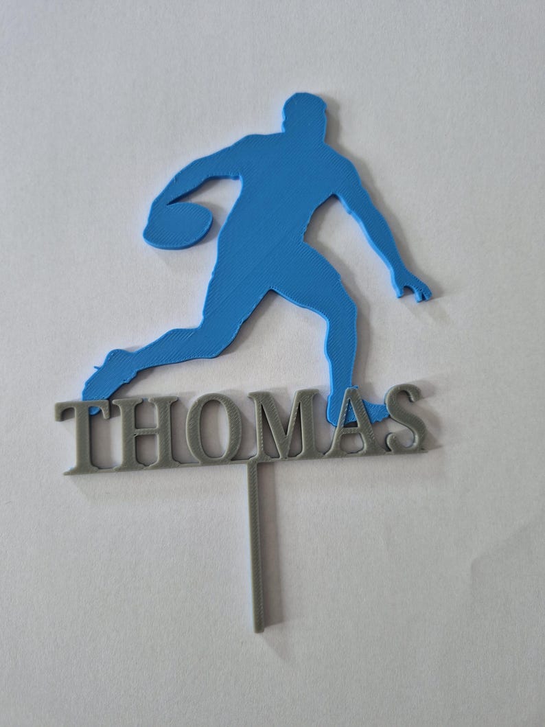 May include: A blue 3D-printed silhouette of a basketball player in motion, positioned above the name THOMAS in grey capital letters. The entire piece is attached to a single vertical grey stake, designed as a decorative cake topper.