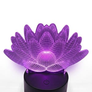 May include: A purple lotus flower-shaped LED lamp. The lamp has a 3D wireframe design and sits on a black base. The lamp is illuminated with a soft purple glow, creating a calming ambiance. The base has a power button.