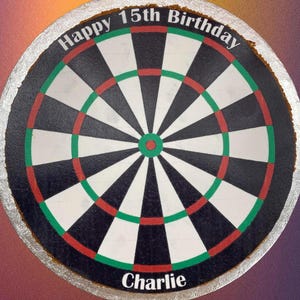 May include: A round cake decorated to look like a dartboard. The cake is white with black and red rings and a green outer ring. The words "Happy 15th Birthday" are written in white on the top of the cake. The name "Charlie" is written in white on the bottom of the cake.