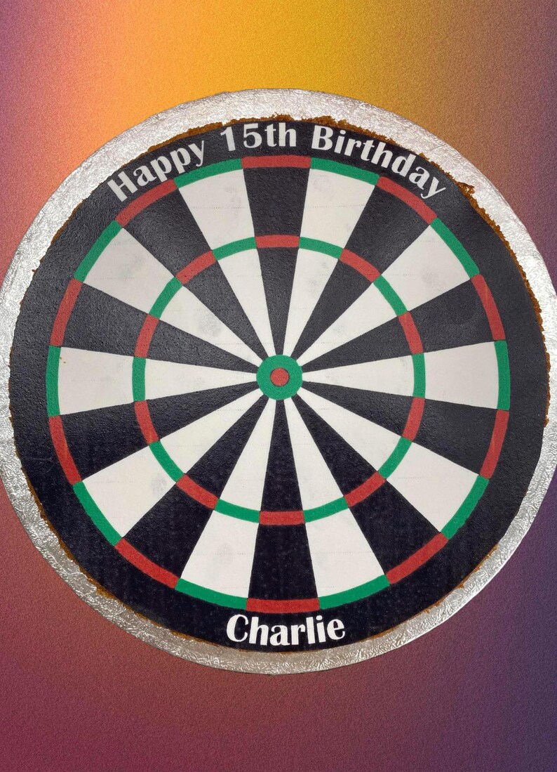 May include: A round cake decorated to look like a dartboard. The cake is white with black and red rings and a green outer ring. The words "Happy 15th Birthday" are written in white on the top of the cake. The name "Charlie" is written in white on the bottom of the cake.