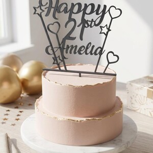Puede incluir: Un pastel rosa de dos pisos con bordes dorados, coronado con un adorno negro que dice "Happy 21st Amelia". El pastel est&aacute; sobre un soporte de m&aacute;rmol blanco. Globos dorados y confeti de estrellas en el fondo.
