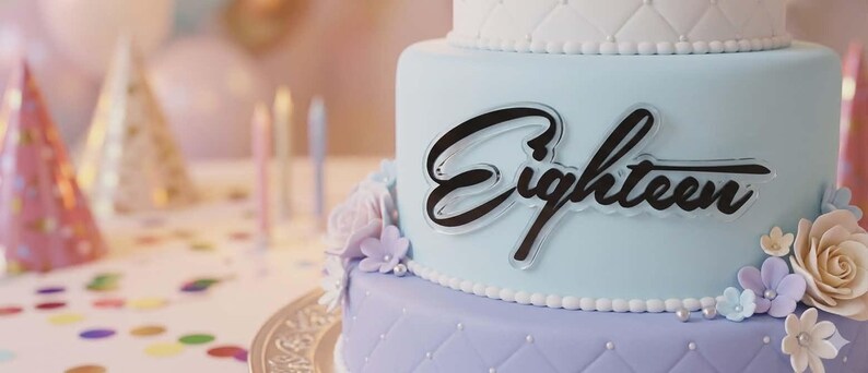 May include: A three-tiered birthday cake with light blue and lavender frosting. The cake is decorated with the word "Eighteen" in black script, and adorned with flowers. Party hats, balloons, and candles are in the background.