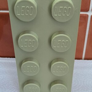 May include: A light green, rectangular container resembling a LEGO brick. The container has a cylindrical opening at the top and features raised LEGO studs on the sides, each embossed with the LEGO logo. The item is designed to hold items.