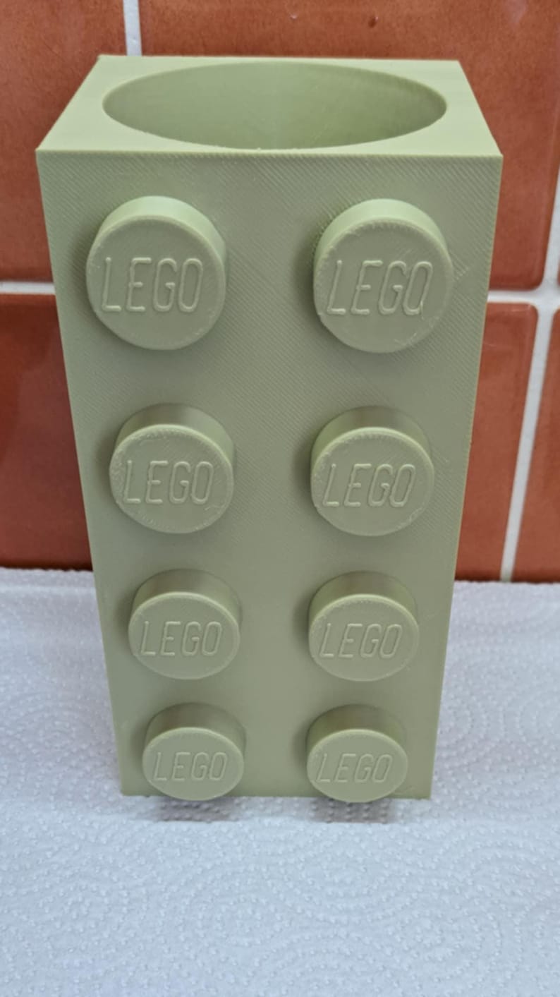 May include: A light green, rectangular container resembling a LEGO brick. The container has a cylindrical opening at the top and features raised LEGO studs on the sides, each embossed with the LEGO logo. The item is designed to hold items.