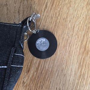 May include: A black denim bag featuring a circular keychain accessory shaped like a vinyl record. The record has a central label with black and white imagery and the text "Back in the Bar" printed in a stylized font, attached by a silver metal ring.