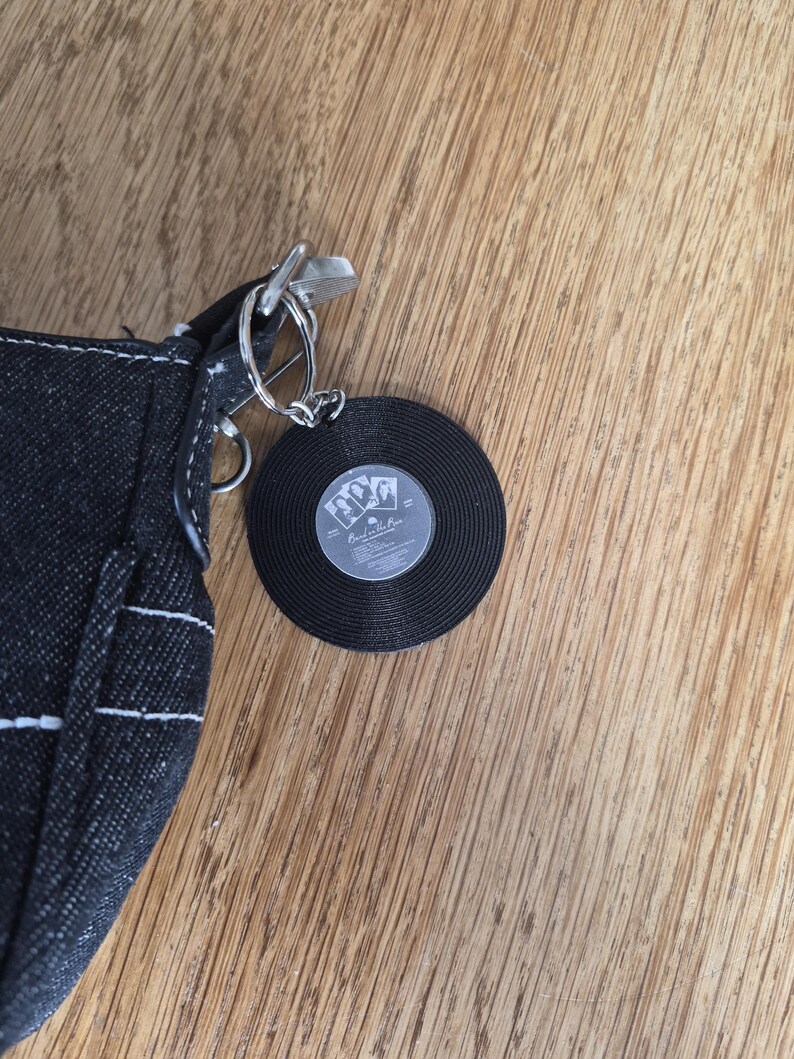 May include: A black denim bag featuring a circular keychain accessory shaped like a vinyl record. The record has a central label with black and white imagery and the text "Back in the Bar" printed in a stylized font, attached by a silver metal ring.