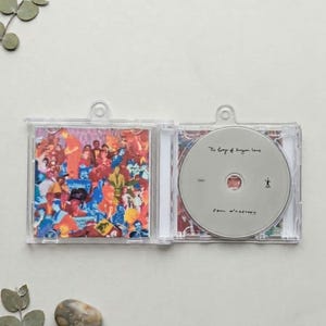 May include: A clear plastic CD case opened to reveal a colourful collage of people on the left panel and a silver disc on the right. The disc features the text The Egypt Station and Paul McCartney in black ink, along with a small stick figure icon.
