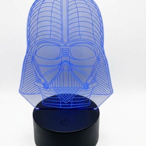 May include: A blue-lighted Darth Vader head-shaped lamp. The lamp features a detailed outline of the character's helmet and mask, with a black base. The light emanates from the base, illuminating the blue lines of the design.