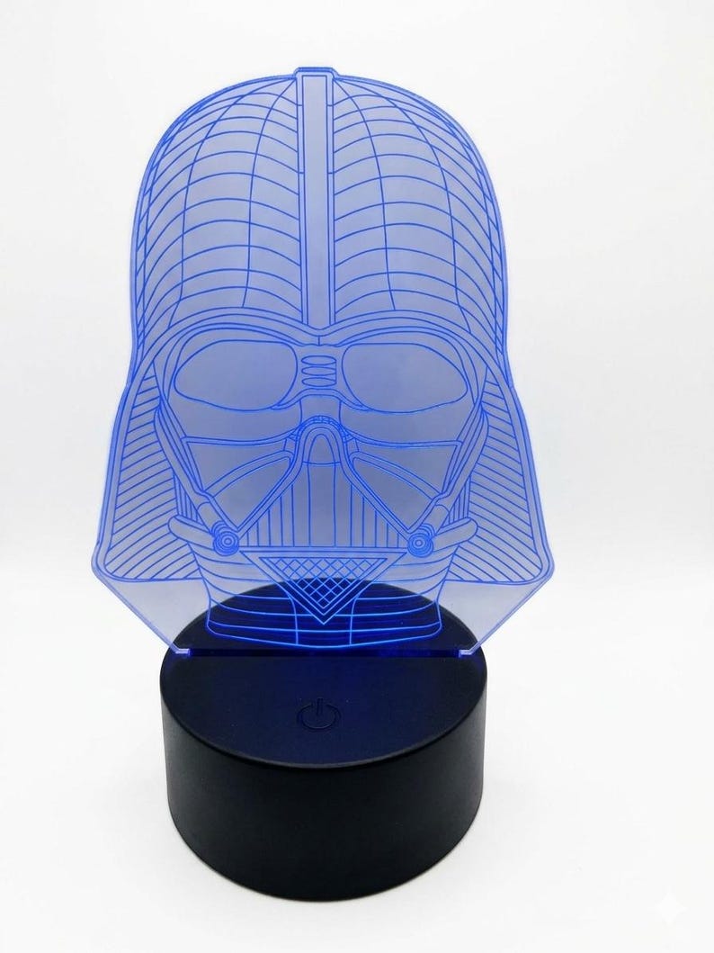 May include: A blue-lighted Darth Vader head-shaped lamp. The lamp features a detailed outline of the character's helmet and mask, with a black base. The light emanates from the base, illuminating the blue lines of the design.