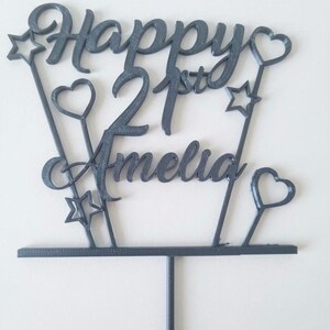 Puede incluir: Un adorno para tarta gris oscuro con las palabras "Happy 21st Amelia" en una fuente cursiva. El dise&ntilde;o incluye estrellas y corazones en varillas delgadas, con una base y un tallo para insertar en un pastel.