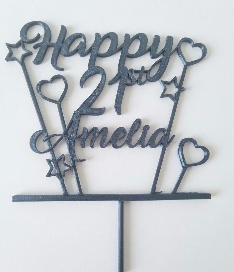 Puede incluir: Un adorno para tarta gris oscuro con las palabras "Happy 21st Amelia" en una fuente cursiva. El dise&ntilde;o incluye estrellas y corazones en varillas delgadas, con una base y un tallo para insertar en un pastel.
