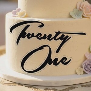 May include: A two-tiered cake with a light beige frosting. The words "Twenty One" are displayed in black script. The cake is decorated with pastel-colored floral accents, including pink, purple, and green roses.
