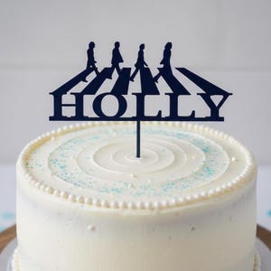 May include: A navy blue cake topper depicting the Abbey Road crossing with four figures. The word "HOLLY" is cut out below the figures. The topper is on a white frosted cake with a pearl border and blue sprinkles.