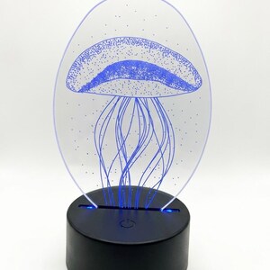 May include: A blue jellyfish design is etched into a clear acrylic panel, illuminated by a black base. The jellyfish has a dotted pattern on its bell and long, flowing tentacles. The base has a power button and emits a soft blue glow.