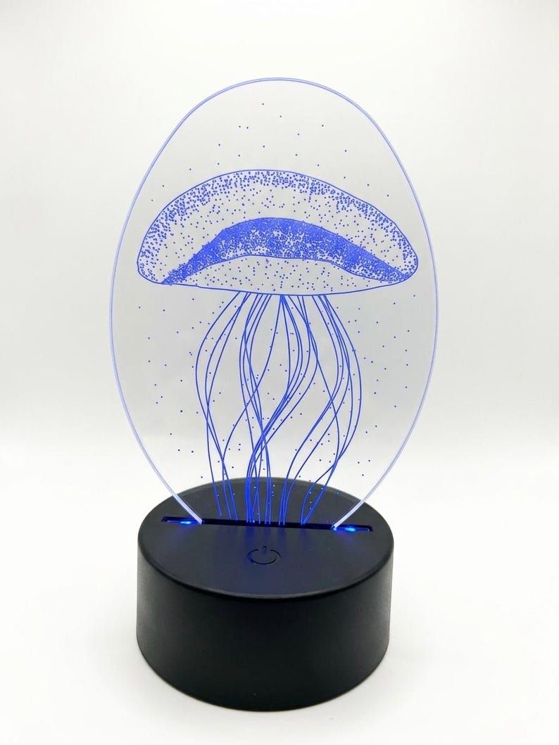 May include: A blue jellyfish design is etched into a clear acrylic panel, illuminated by a black base. The jellyfish has a dotted pattern on its bell and long, flowing tentacles. The base has a power button and emits a soft blue glow.