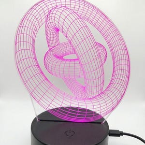 May include: A pink 3D-printed geometric sculpture illuminated by an LED light. The sculpture features a complex, interwoven design, mounted on a black base with a power button. A USB cable extends from the base.