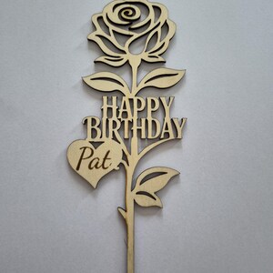 May include: A wooden cake topper in the shape of a rose with the words "HAPPY BIRTHDAY Pat" cut out. The topper is made of light-colored wood and features a detailed rose blossom, leaves, and a heart shape.