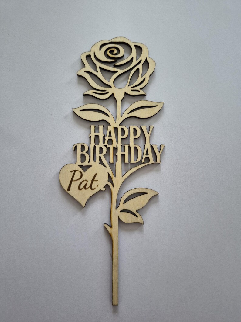 May include: A wooden cake topper in the shape of a rose with the words "HAPPY BIRTHDAY Pat" cut out. The topper is made of light-colored wood and features a detailed rose blossom, leaves, and a heart shape.
