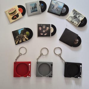 May include: A collection of miniature vinyl record keychains featuring iconic album covers including The Beatles, Queen, and The Rolling Stones, alongside three square plastic record player frames in red, grey, and black with metal key rings.