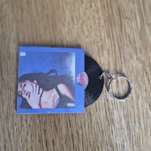May include: A keychain featuring a miniature black vinyl record partially inside a square blue sleeve. The sleeve displays a photographic print of a person with dark hair and a black top, with a small white square and an advisory label visible.