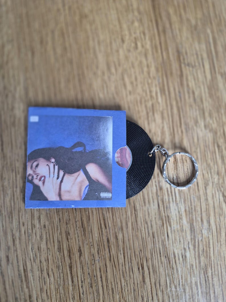 May include: A keychain featuring a miniature black vinyl record partially inside a square blue sleeve. The sleeve displays a photographic print of a person with dark hair and a black top, with a small white square and an advisory label visible.