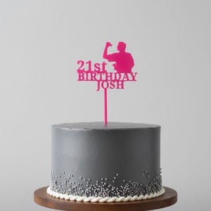 May include: A gray frosted cake with a pink cake topper that reads "21st BIRTHDAY JOSH". The cake is on a wooden cake stand and is decorated with silver sprinkles along the bottom edge.