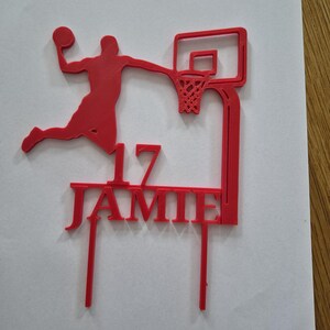 May include: A red cake topper featuring a silhouette of a basketball player dunking a ball into a hoop. The design includes the number 17 and the name JAMIE in capital letters, supported by two vertical prongs at the base for insertion into a cake.