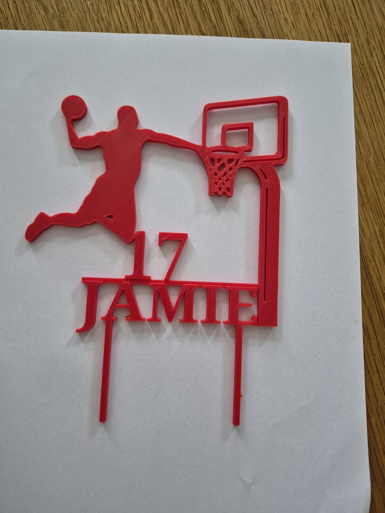 May include: A red cake topper featuring a silhouette of a basketball player dunking a ball into a hoop. The design includes the number 17 and the name JAMIE in capital letters, supported by two vertical prongs at the base for insertion into a cake.
