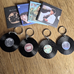 May include: Four circular black vinyl record keychains with central album art labels, accompanied by four corresponding square album cover prints. The labels feature various musical imagery, including a group silhouette, teeth, and text.