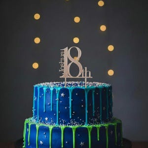 May include: A two-tiered birthday cake with blue frosting and green and blue drips. The cake is decorated with silver sprinkles and star-shaped sprinkles. A wooden cake topper reads "Joshua 18th".
