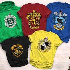 May include: A collection of five shirts and a hoodie featuring Harry Potter house crests. The green hoodie displays Slytherin, the red shirt Gryffindor, the blue shirt Ravenclaw, the yellow shirt Hufflepuff, and the black shirt the Hogwarts crest.