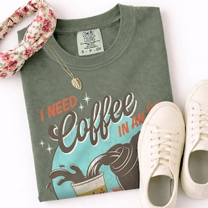 May include: An olive green t-shirt with the text "I NEED Coffee IN AN" in a retro design. The shirt is paired with white sneakers, a floral headband, and a gold leaf pendant necklace. The shirt has a Comfort Colors tag.