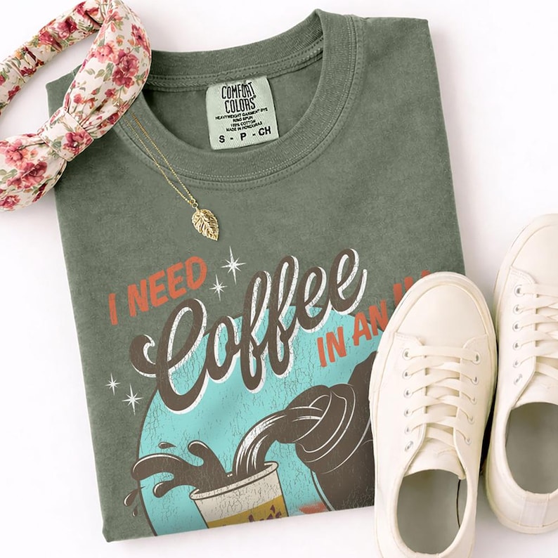 May include: An olive green t-shirt with the text "I NEED Coffee IN AN" in a retro design. The shirt is paired with white sneakers, a floral headband, and a gold leaf pendant necklace. The shirt has a Comfort Colors tag.