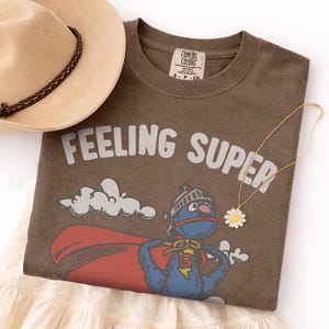 May include: A brown t-shirt with the words "FEELING SUPER" in white. The shirt features a cartoon superhero character with a red cape. A tan hat with a brown band and a daisy pendant necklace are also in the image.