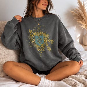 May include: A dark gray sweatshirt featuring a gold and teal crest design. The crest has floral accents and a letter "B" in the center. The sweatshirt is a relaxed fit and has a vintage, washed appearance.