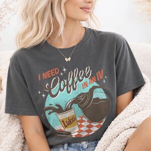 May include: Dark gray t-shirt with the text "I NEED Coffee IN AN IV" in a retro design. The graphic shows coffee pouring into a cup labeled "Luke's" with a red and white checkered background. A fun, coffee-themed tee.