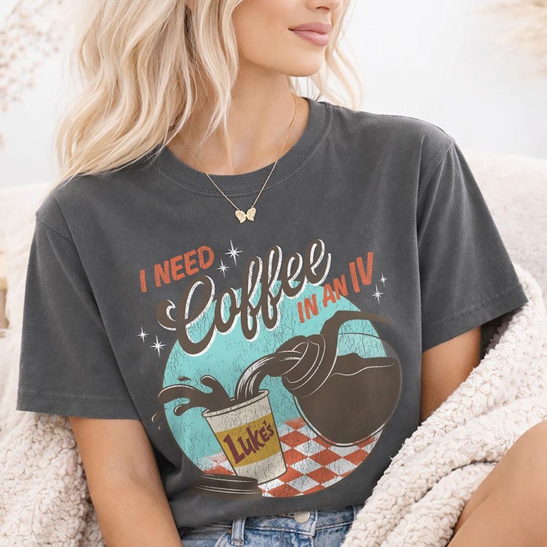 May include: Dark gray t-shirt with the text "I NEED Coffee IN AN IV" in a retro design. The graphic shows coffee pouring into a cup labeled "Luke's" with a red and white checkered background. A fun, coffee-themed tee.