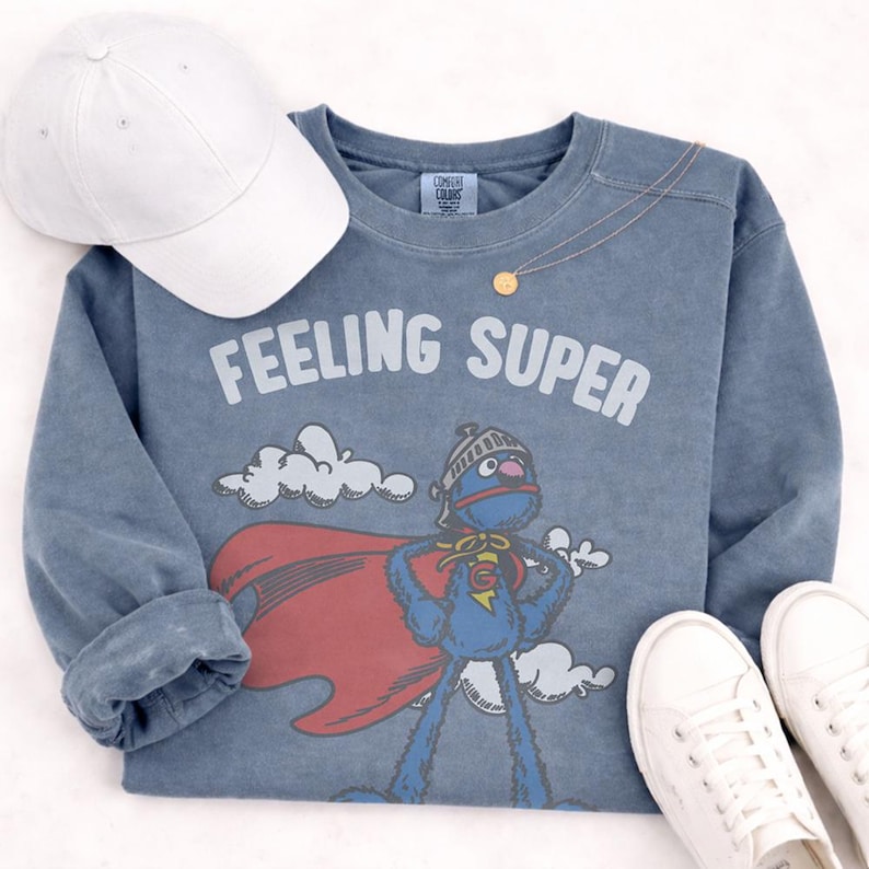 May include: A blue long-sleeve sweatshirt with the words "FEELING SUPER" in white. The graphic features a blue cartoon character with a red cape. A white baseball cap and white sneakers are also in the image.