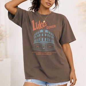 Op de afbeelding: Bruin t-shirt met de tekst "Luke's Diner" in oranje en een blauwe illustratie van een dinergebouw. Het shirt bevat ook de tekst "Breakfast, Lunch & Dinner - No Cell Phones - Stars Hollow, CT".
