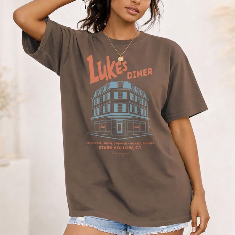 Op de afbeelding: Bruin t-shirt met de tekst "Luke's Diner" in oranje en een blauwe illustratie van een dinergebouw. Het shirt bevat ook de tekst "Breakfast, Lunch & Dinner - No Cell Phones - Stars Hollow, CT".