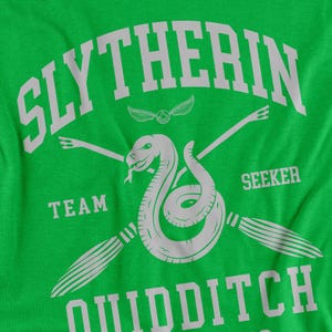 May include: Bright green t-shirt with a white graphic design. The design features the word "SLYTHERIN" arched across the top, a snake, crossed brooms, and the words "TEAM", "SEEKER", and "QUIDDITCH".